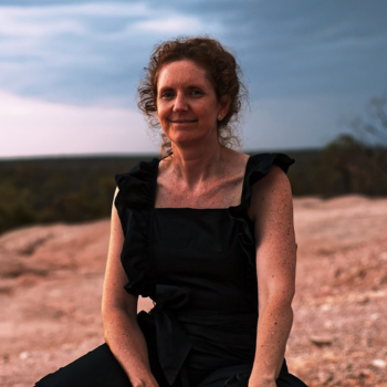 A woman sitting outdoors at sunset, wearing a black dress, with a soft landscape background.